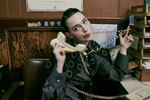 Stylish woman in a grey suit sits at a desk and poses with a vintage telephone in an indoor office environment with warm lighting.