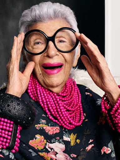 Elderly woman with round glasses, floral shirt, and pink beaded necklace smiles at the camera, placing her hands near her face.