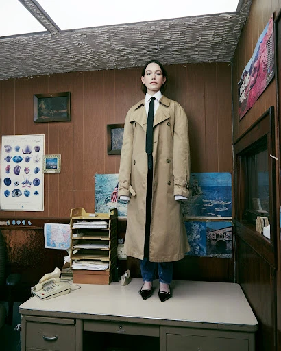 A model stands on a desk in an office wearing a trench coat, tie, and heels against a wood-paneled wall with various posters and artwork.