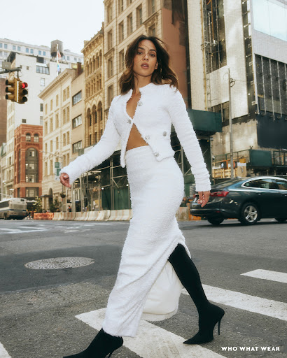 A woman in a white two-piece set crosses a city street, showcasing a coordinated jacket and skirt with black tights and heeled boots.