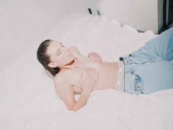 A topless woman lying on a white bed wearing light blue jeans with visible branded waistband, styled by Anna Katsanis.