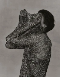 Model wearing a metallic chainmail garment held up over the face against a neutral textured background