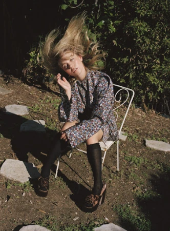 Woman in floral print dress and platform shoes sitting on a white metal chair in a sunlit garden, flipping her hair.