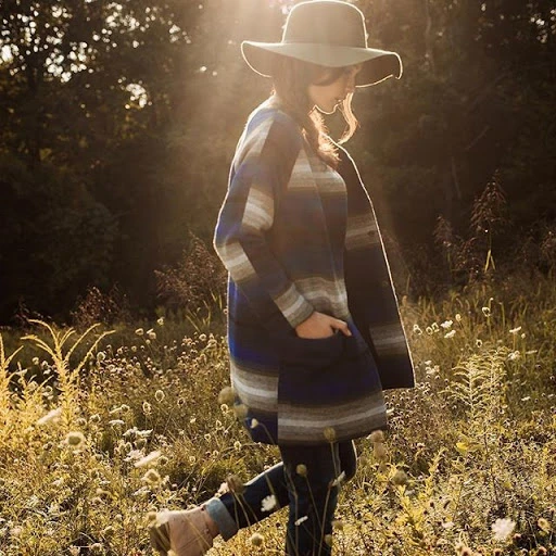 Woman in wide-brimmed hat and patterned coat walking through a field of wildflowers on a sunny day. Fashion shot with natural light.