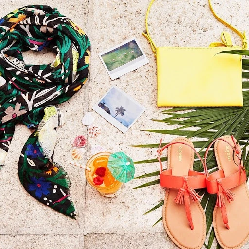 Tropical vacation still life with orange sandals, yellow purse, floral scarf, and fruity drink, set on a stone surface with palm fronds.