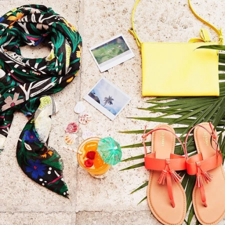 Tropical vacation still life with orange sandals, yellow purse, floral scarf, and fruity drink, set on a stone surface with palm fronds.