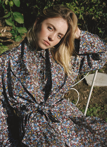 Young woman with blond hair in a floral dress sitting in garden, posing for a portrait in natural light.