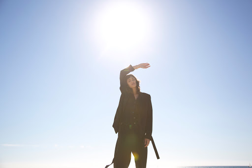 A woman in a dark suit poses outdoors against a bright, clear blue sky with the sun shining behind her, obscuring her facial details.