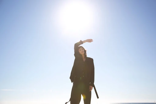 A woman in a dark suit poses outdoors against a bright, clear blue sky with the sun shining behind her, obscuring her facial details.