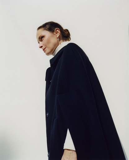 Woman in a dark blue cape and white turtleneck looking up in a bright indoor setting, captured from a low angle.