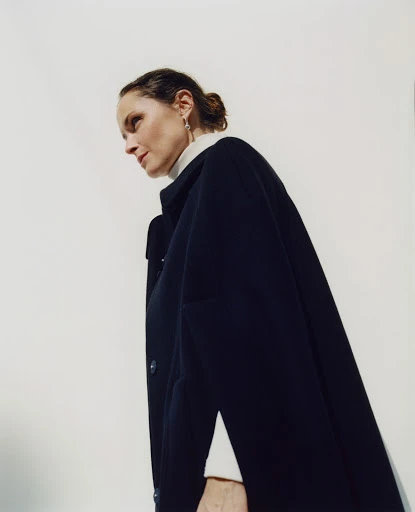 Woman in a dark blue cape and white turtleneck looking up in a bright indoor setting, captured from a low angle.
