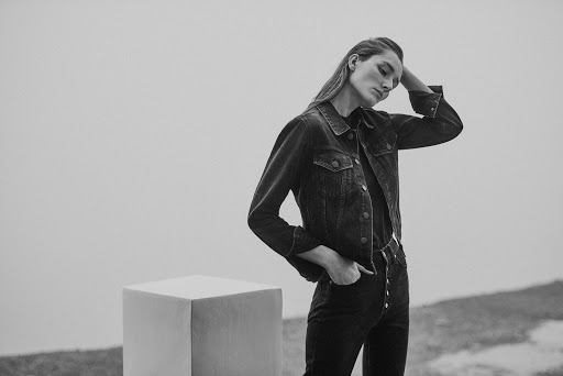 Monochrome fashion portrait of a model in a dark denim jacket and matching jeans posing next to a white block prop.