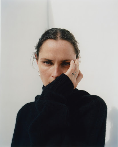 Portrait of an adult female with hand near face, wearing a black turtleneck, with neutral facial expression against a white wall.