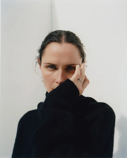 Portrait of an adult female with hand near face, wearing a black turtleneck, with neutral facial expression against a white wall.