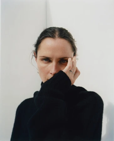 Portrait of an adult female with hand near face, wearing a black turtleneck, with neutral facial expression against a white wall.