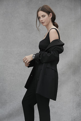 Portrait of a brunette woman in a black top and pants, with a black jacket draped over her shoulders, posing against a grey background.