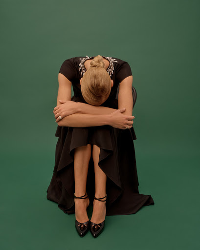 Woman in a black dress and heels, seated with head down against a green background. Fashion shot with elegant and minimalist composition.