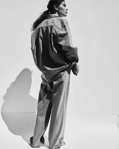 A woman in a denim shirt and jeans poses in a studio with her back to the viewer. The black and white image is brightly lit.