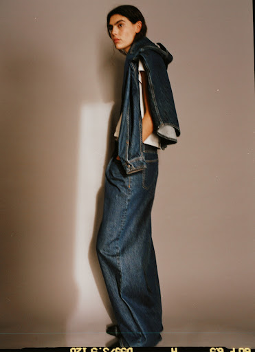 Fashion shot of a woman wearing a denim jacket and jeans, posed in a photo studio with a neutral background and studio lighting.