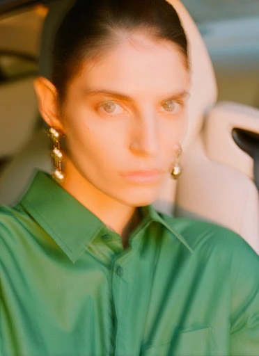 A woman with dark hair and green eyes wearing a green collared shirt and gold earrings is posed in front of a tan car seat.