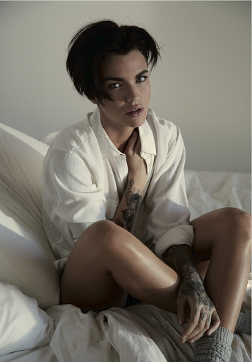 Ruby Rose with dark short hair wearing a white collared shirt and grey socks seated on a bed with white pillows.