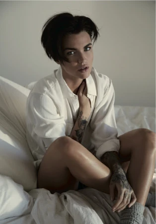 Ruby Rose with dark short hair wearing a white collared shirt and grey socks seated on a bed with white pillows.