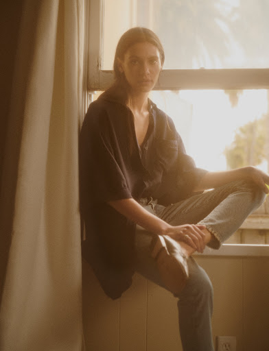 A young woman with light skin and brown hair sits on a windowsill wearing a dark top and light blue jeans, looking directly at the camera.