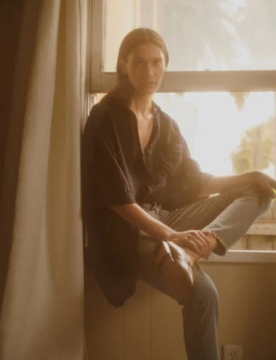 A young woman with light skin and brown hair sits on a windowsill wearing a dark top and light blue jeans, looking directly at the camera.