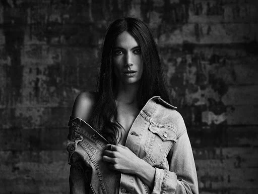 Black and white portrait of a woman wearing an off-the-shoulder denim jacket against a weathered wall background.