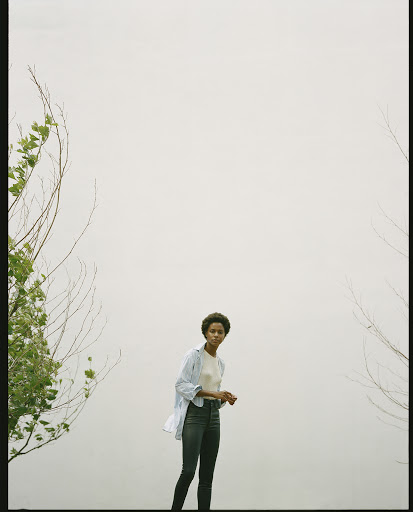 A young African-descent woman stands in front of a washed-out grey background, wearing jeans and a light blue shirt, framed by branches.