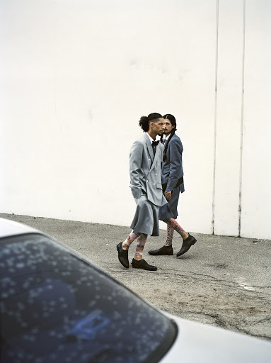 Two men in modern suits walk outdoors next to a white wall with a blurred car foreground. Street fashion editorial photography.