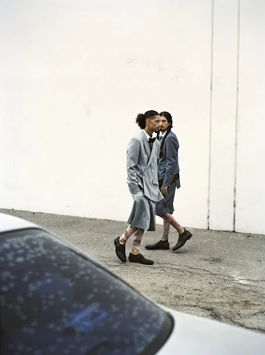 Two men in modern suits walk outdoors next to a white wall with a blurred car foreground. Street fashion editorial photography.