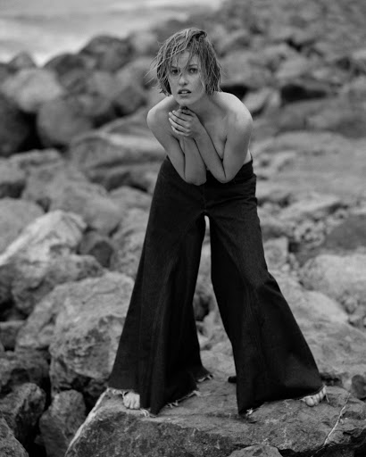 A woman poses on rocks in bell-bottom jeans in a black-and-white outdoor fashion shot. She is standing with her hands clasped.
