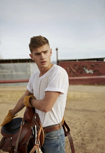 Man in white t-shirt holds brown leather saddle, wearing jeans and tan gloves, in an outdoor setting with a cloudy sky.