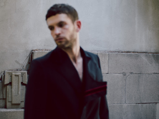 Man in a dark jacket poses outdoors in front of a textured, light-colored wall with geometric ornamentation and natural light.