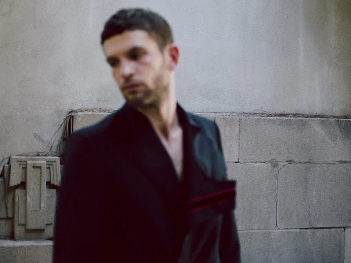 Man in a dark jacket poses outdoors in front of a textured, light-colored wall with geometric ornamentation and natural light.