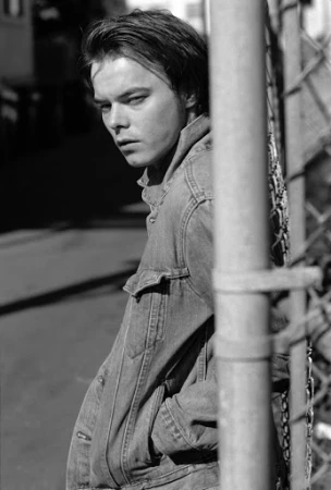 Monochrome portrait of a young man in a denim jacket standing against a chain-link fence, looking back.