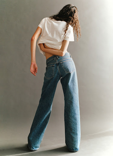 Young woman stands in a photo studio, wearing a white t-shirt and blue jeans, looking back over her shoulder with soft studio lighting.