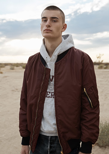 Man in burgundy bomber jacket and grey hoodie stands outdoors in a desert-like landscape with a cloudy sky in a medium shot.