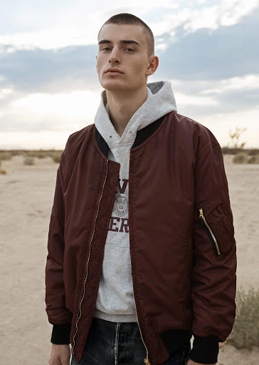 Man in burgundy bomber jacket and grey hoodie stands outdoors in a desert-like landscape with a cloudy sky in a medium shot.