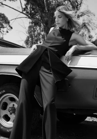 Monochrome fashion photograph of a woman in dark attire leaning against a classic car outdoors.