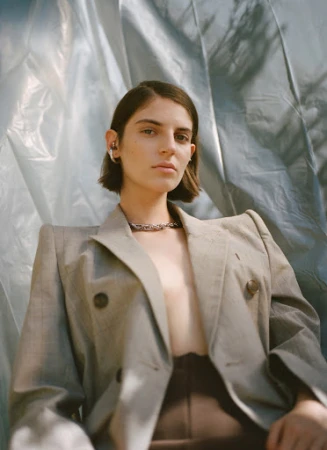Woman in an unbuttoned grey blazer poses in front of a light blue backdrop wearing a chain necklace. Medium shot, portrait orientation.