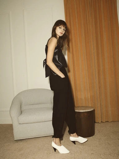 A woman with bangs poses indoors wearing a black top and pants, with white shoes. A chair and side table are visible in the background.