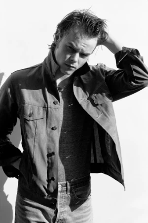 Monochrome portrait of a young man wearing a denim jacket, gray t-shirt and jeans, posing with his hand in his hair against a white wall.
