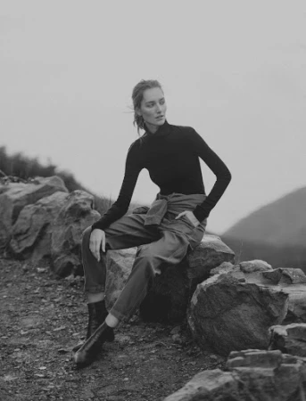Model wearing a black turtleneck and dark trousers seated on rocks in an outdoor landscape styled by Annina Mislin