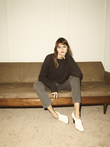 A woman with bangs wearing a grey sweater and jeans sits on a brown couch with white shoes. Neutral, indoor setting with soft lighting.