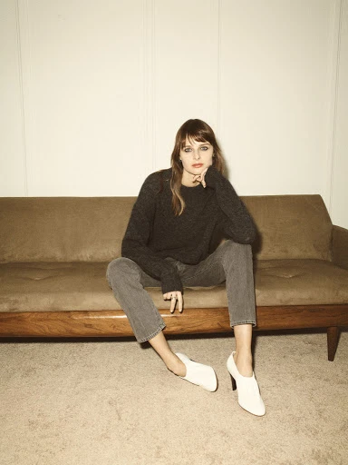 A woman with bangs wearing a grey sweater and jeans sits on a brown couch with white shoes. Neutral, indoor setting with soft lighting.