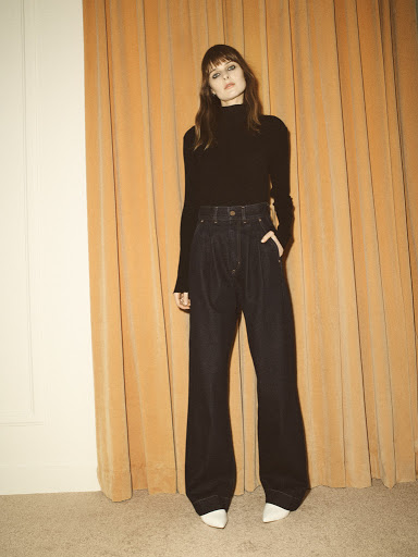 Full-length shot of a slender adult woman posing in wide-leg jeans, a black turtleneck, and white heels in front of a gold curtain.
