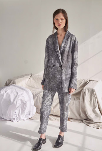 Adult female in a gray patterned pantsuit and black leather shoes standing in front of a white backdrop with draped fabric.
