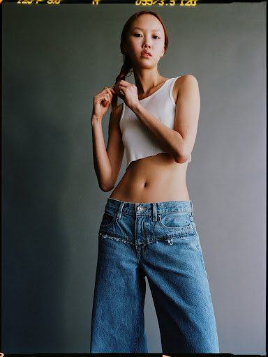 Adult woman in a white tank top and blue denim wide-leg jeans stands in a studio with a gray background, posing for a fashion shot.